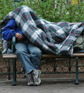 person experiencing homelessness, on park bench with blanket over head